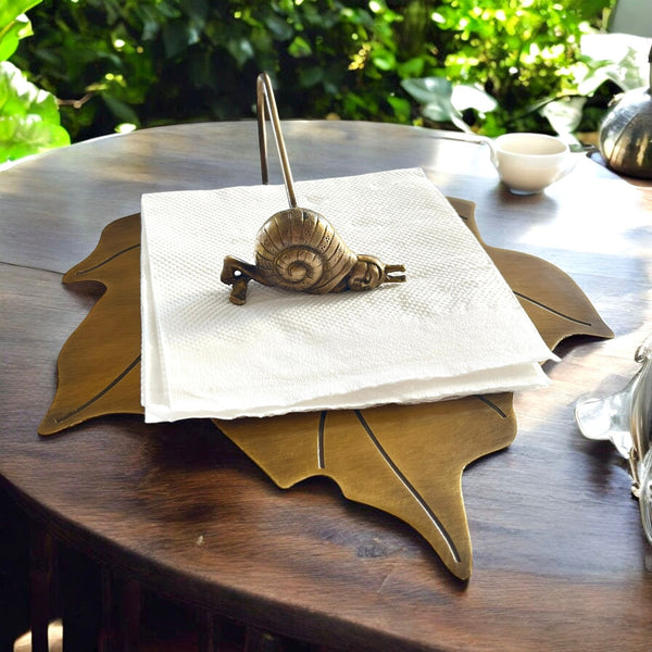 Grace Napkin Holder featuring a golden snail design on a leaf-shaped base, elegantly holding a stack of white napkins on a wooden table, with natural foliage in the background.